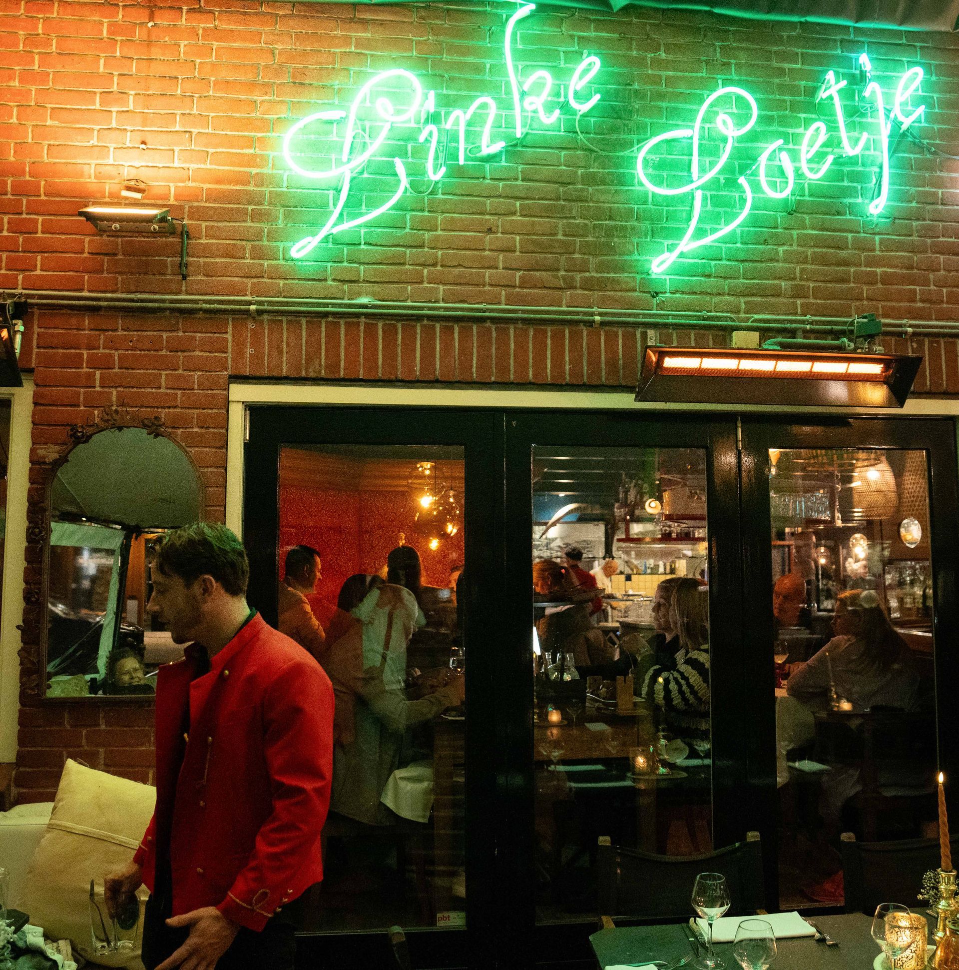 Een man in een rood jasje staat voor een neonbord met de tekst linke boeije. . Bij Linke Loetje