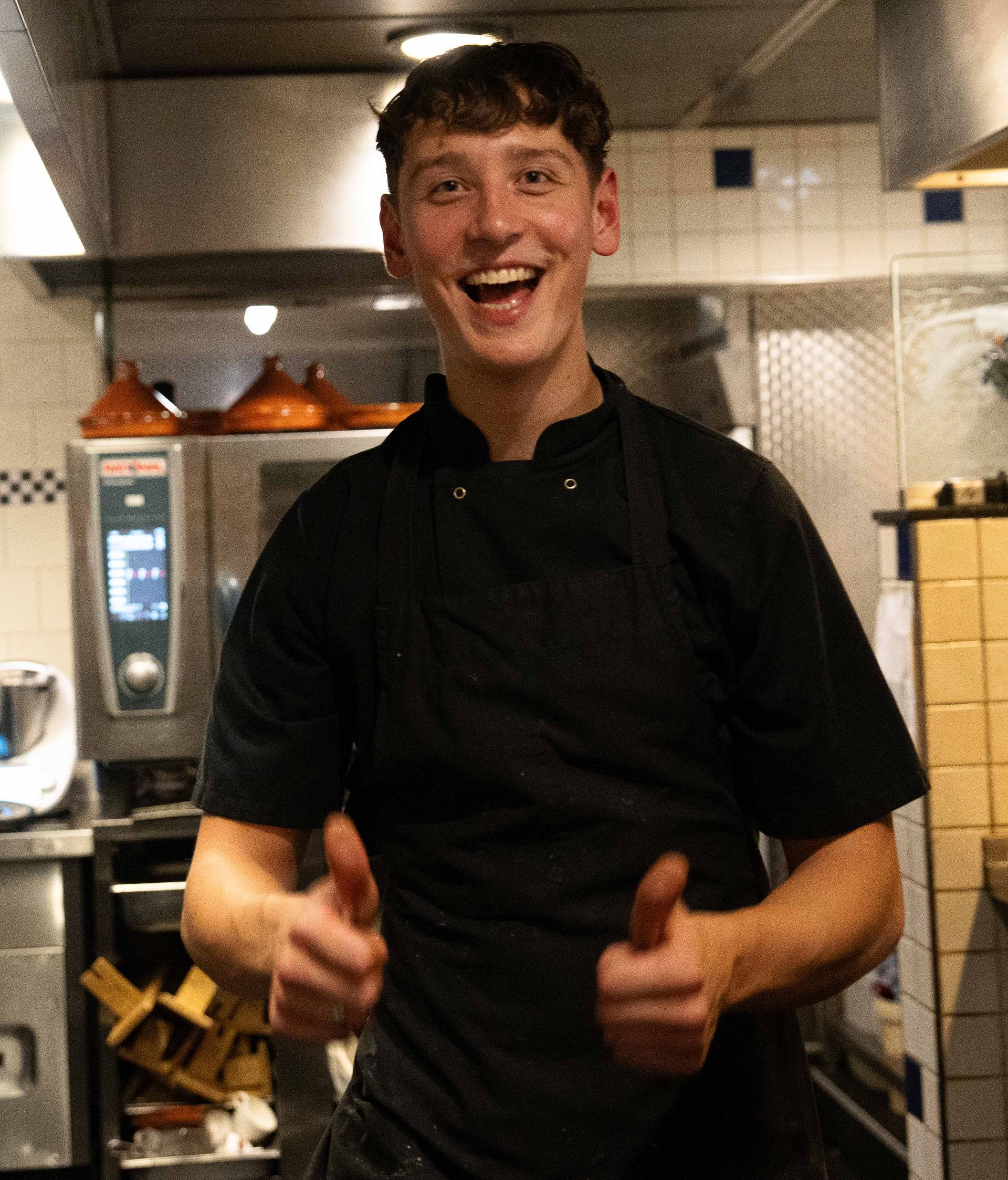 Een man in een zwart overhemd en schort steekt zijn duim omhoog. Bij Linke Loetje