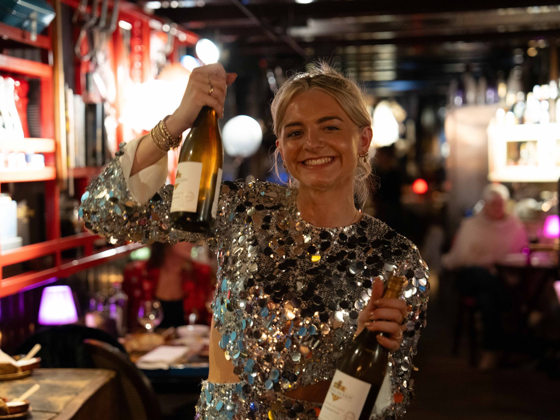 Een vrouw in een jurk met pailletten houdt een fles wijn vast.