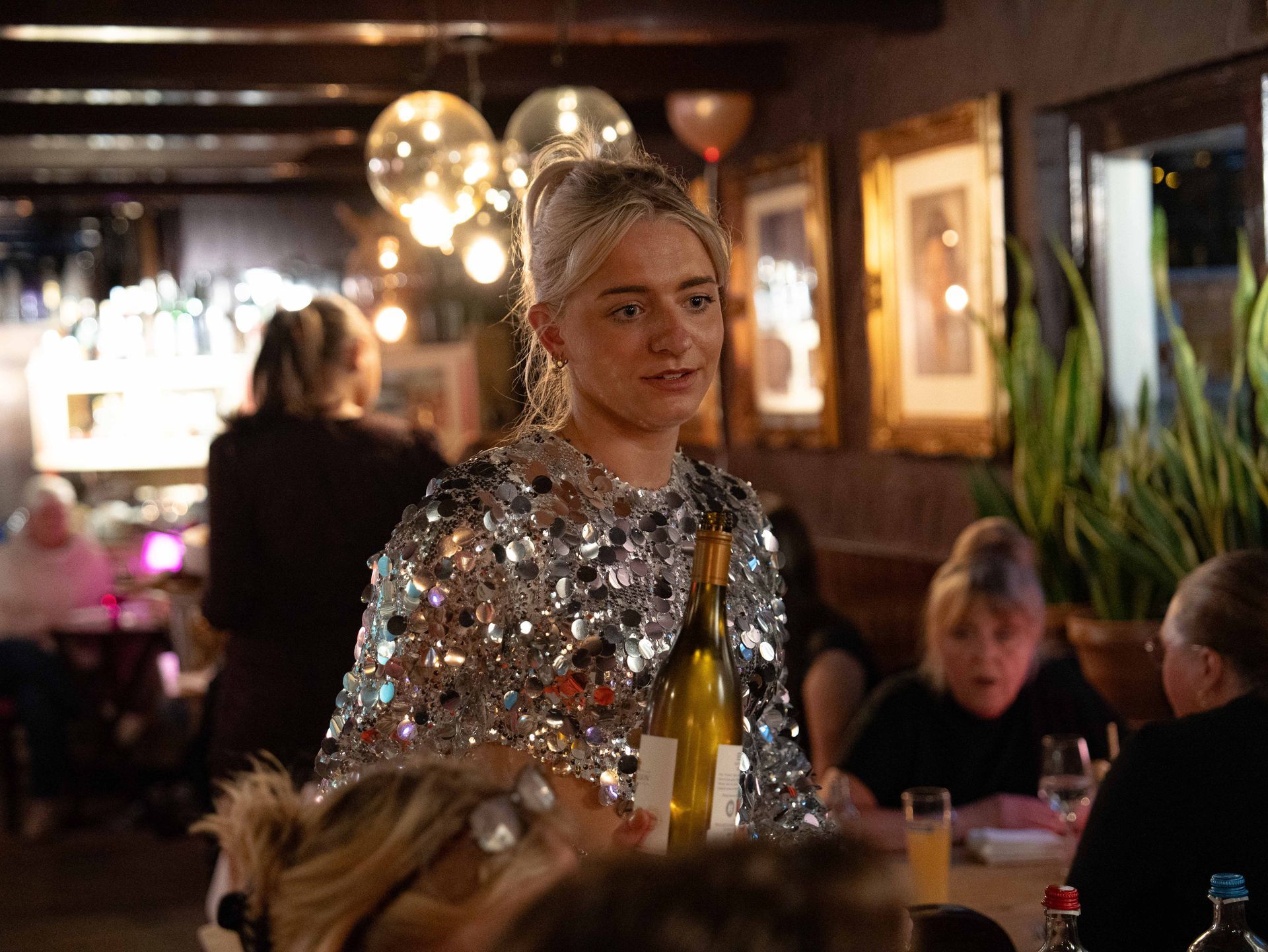 Een vrouw in een jurk met pailletten houdt een fles wijn vast in een restaurant.