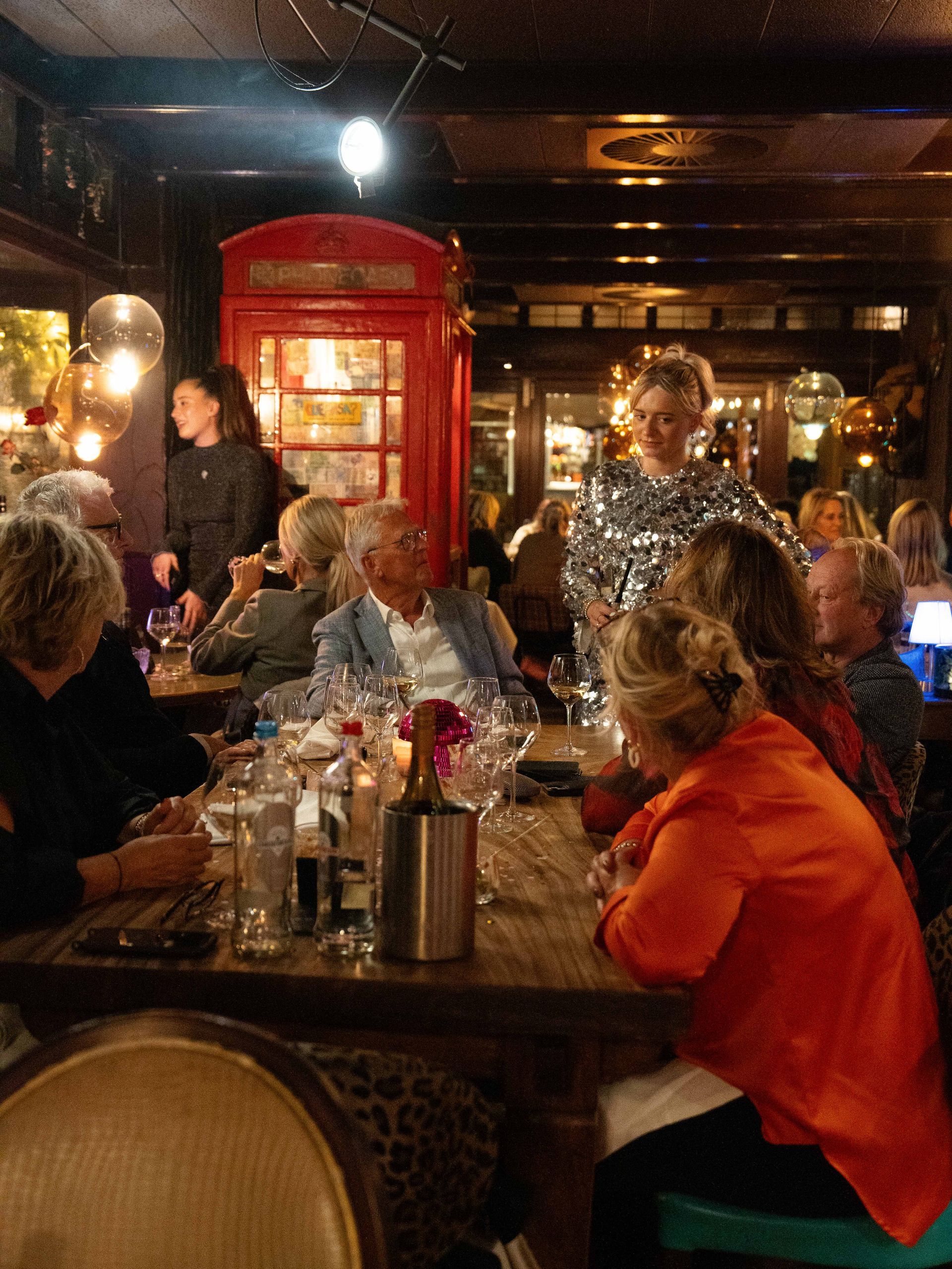 Een groep mensen zit aan tafels in een restaurant met een rode telefooncel op  . Bij Linke Loetjede achtergrond
