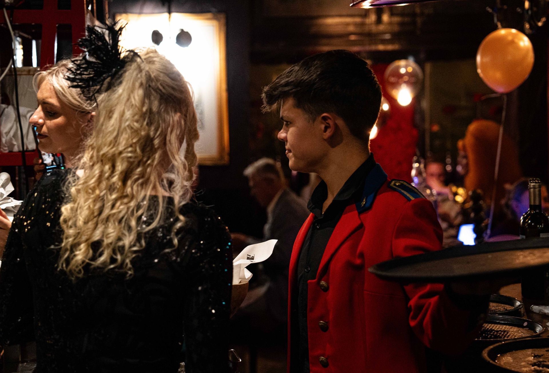 Een man in een rood jasje houdt een dienblad vast naast een vrouw in een zwarte jurk.