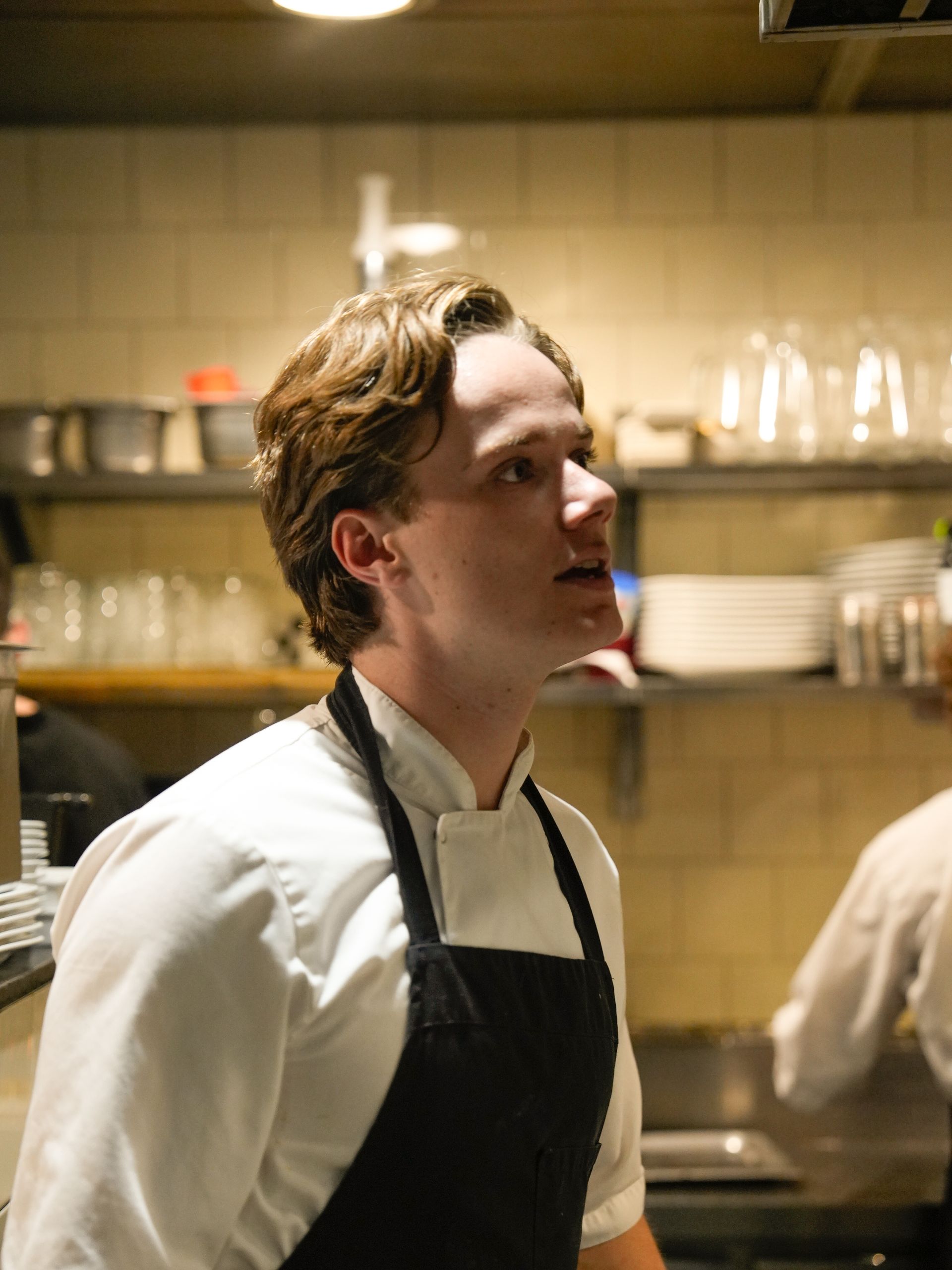 Een man in een wit overhemd en een zwart schort staat in een keuken. Bij Linke Loetje