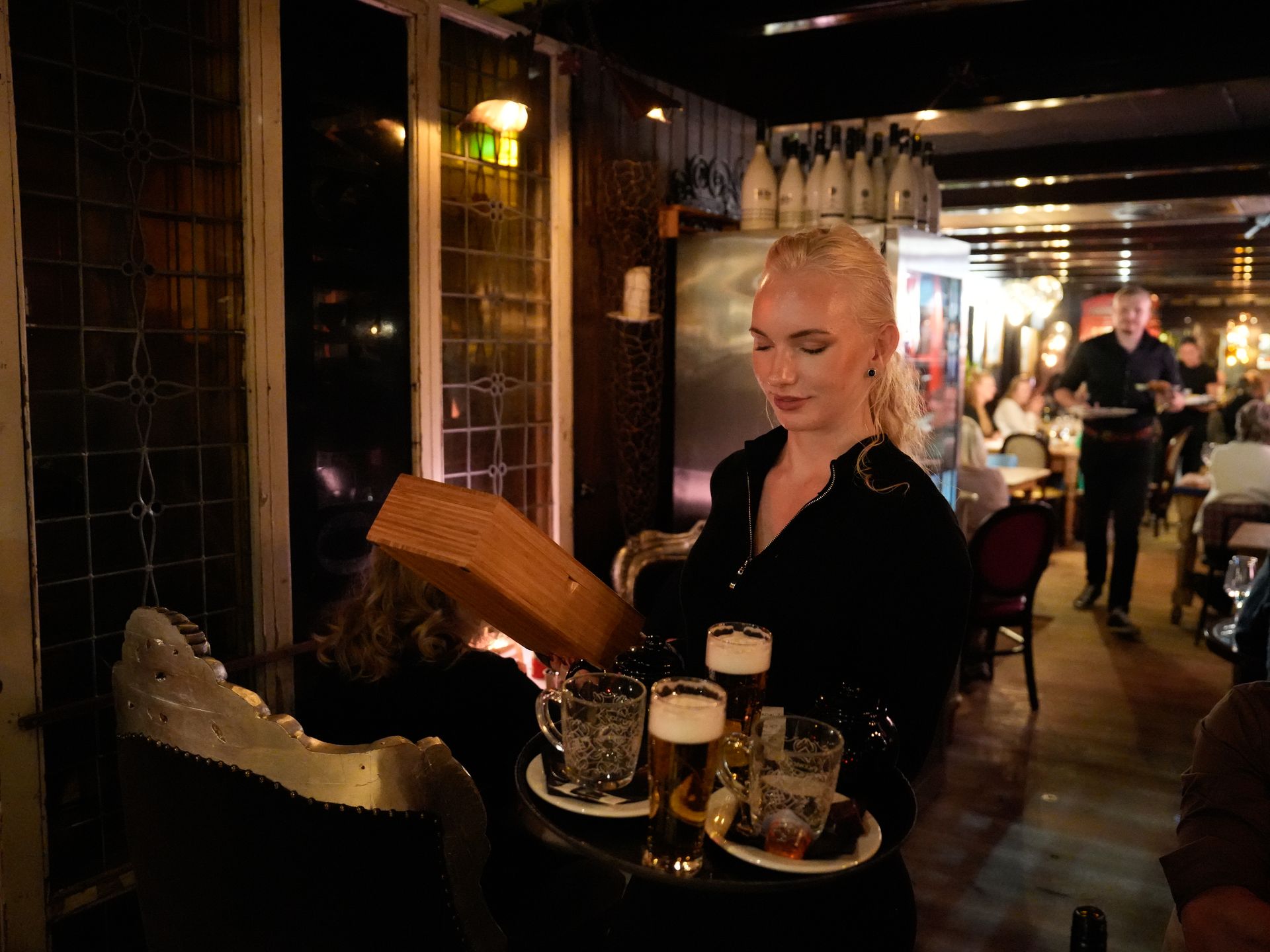 Een vrouw houdt een dienblad met bier en een menukaart vast in een restaurant. . Bij Linke Loetje