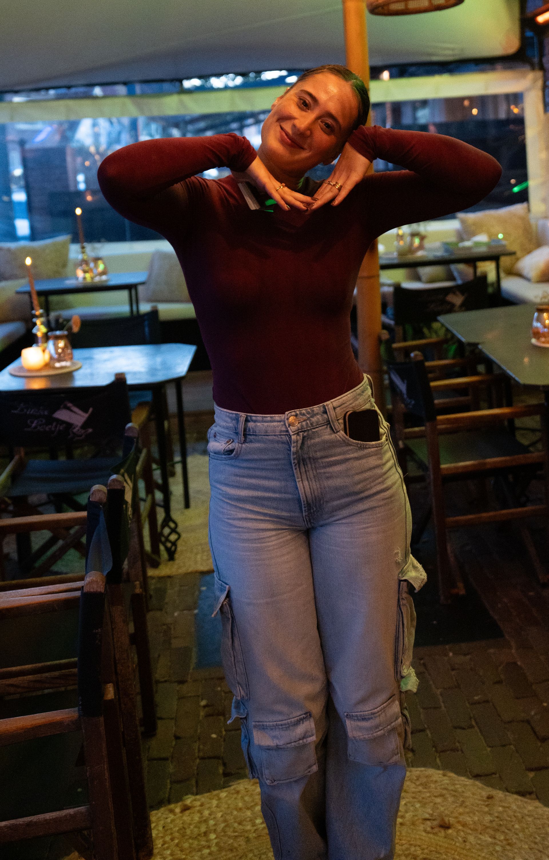 Een vrouw in een rode trui en jeans staat in een restaurant met haar handen achter haar hoofd.. Bij Linke Loetje