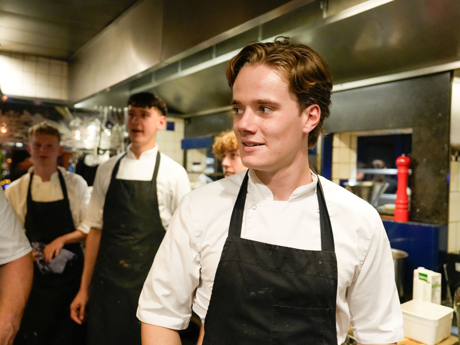 Een man met een schort staat in een keuken met andere koks.. Bij Linke Loetje
