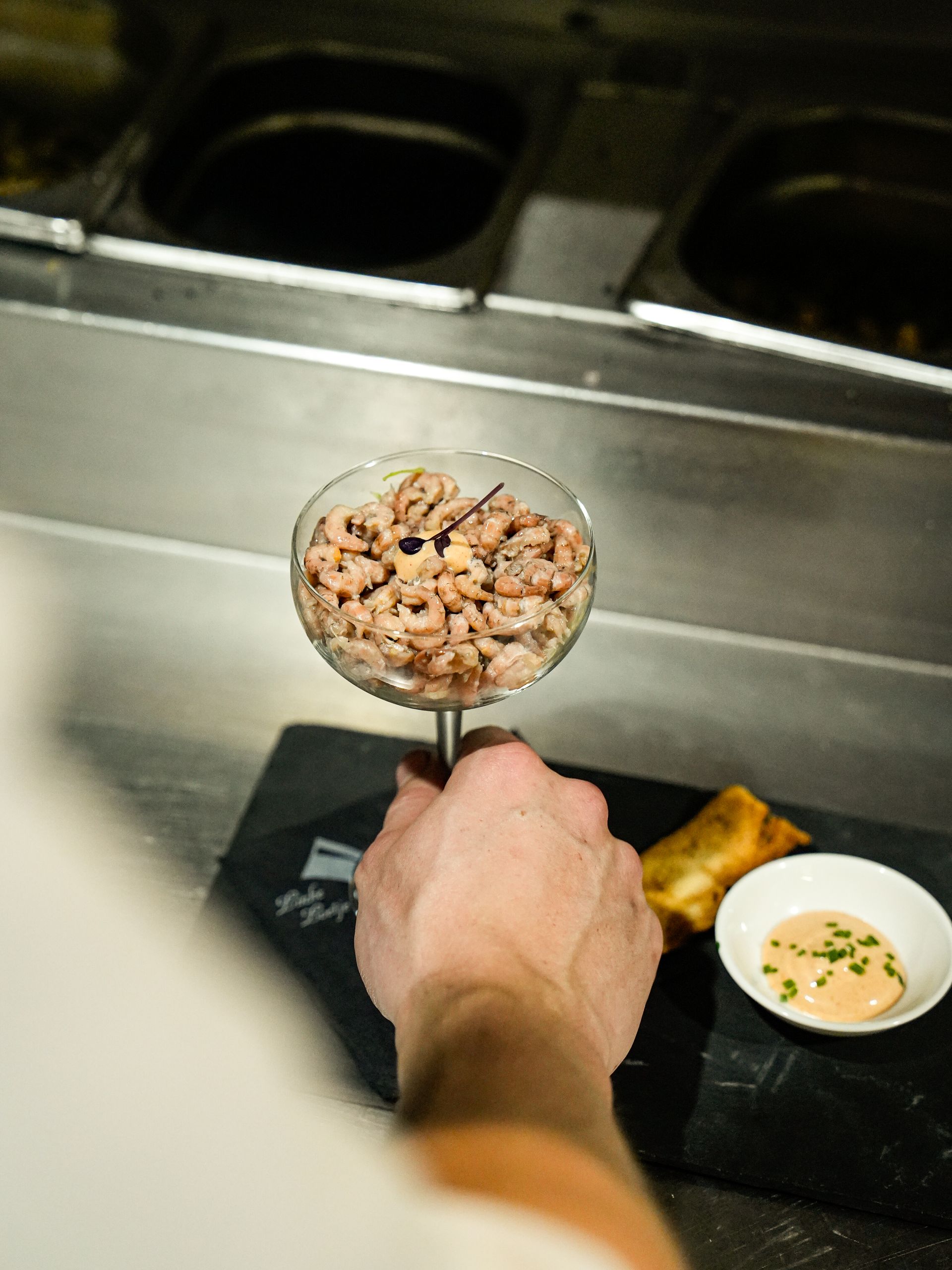 Een persoon houdt een glas vast gevuld met garnalen op een stokje.. Bij Linke Loetje