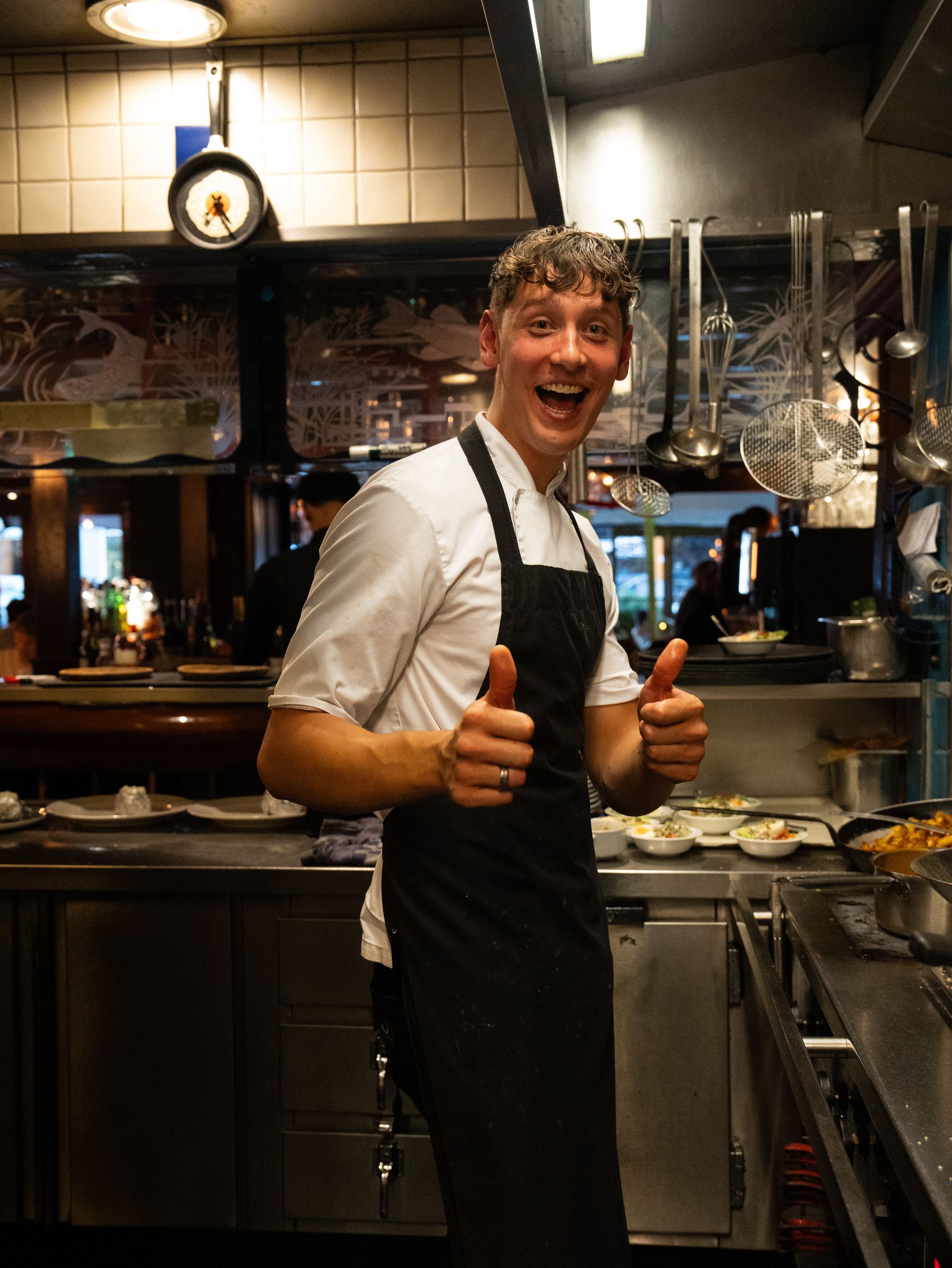 Een man met een schort steekt zijn duim omhoog in een keuken. Bij Linke Loetje
