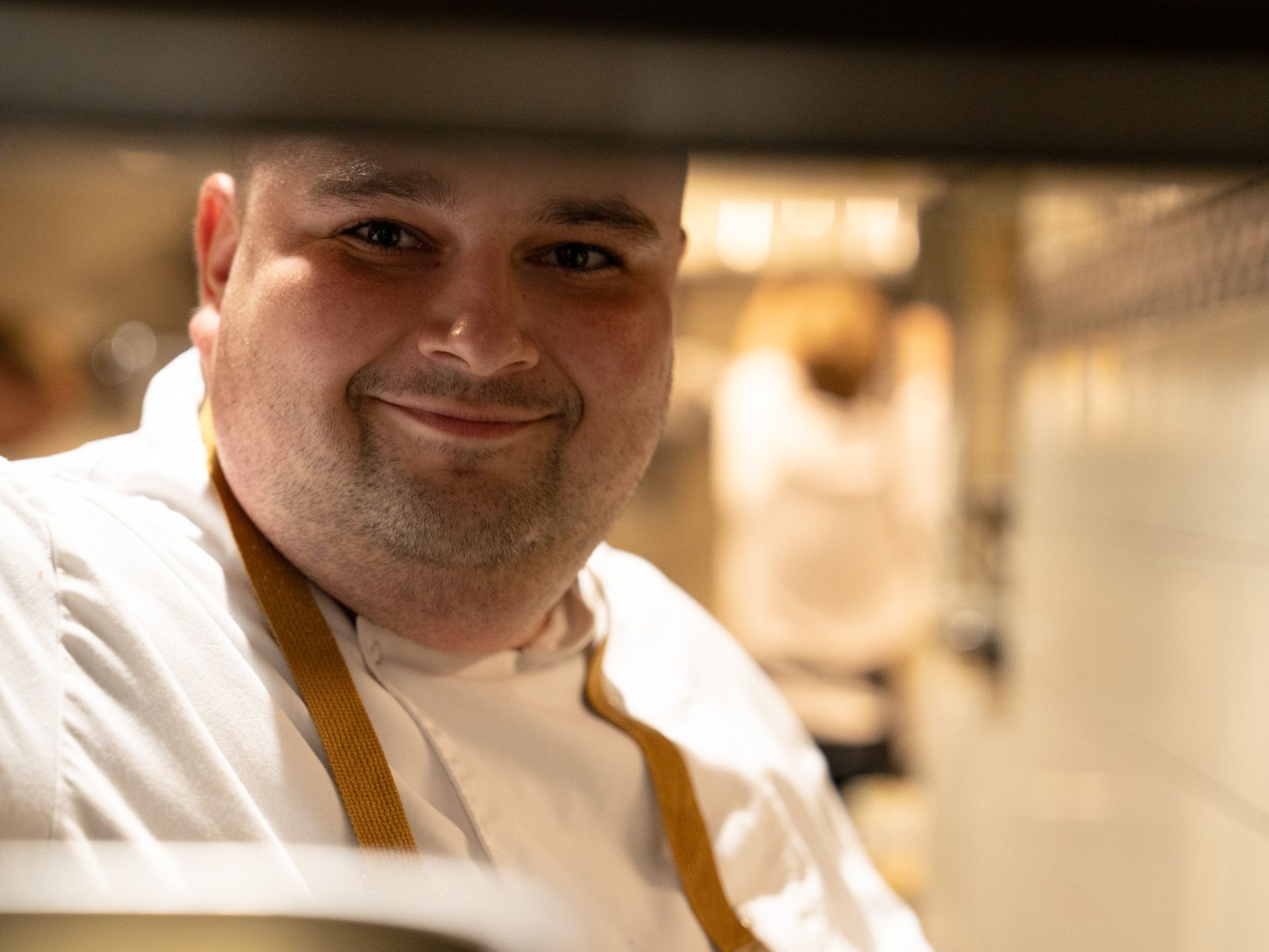 Een man in een wit overhemd en een schort glimlacht in de keuken. . Bij Linke Loetje