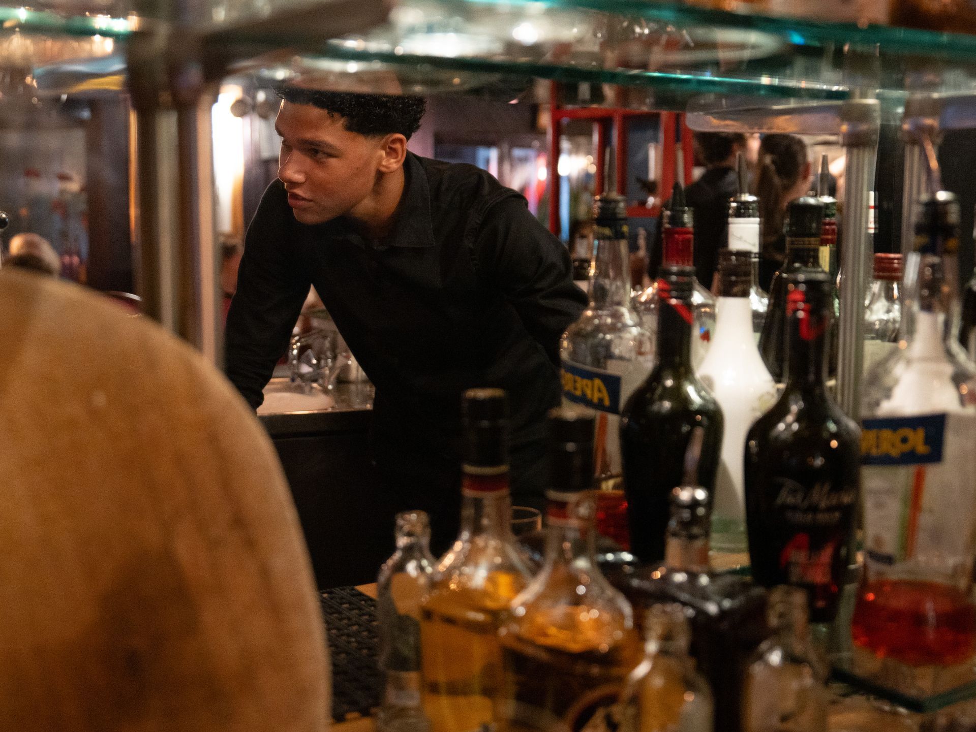 Een barman staat achter een glazen plank gevuld met flessen alcohol. Bij Linke Loetje