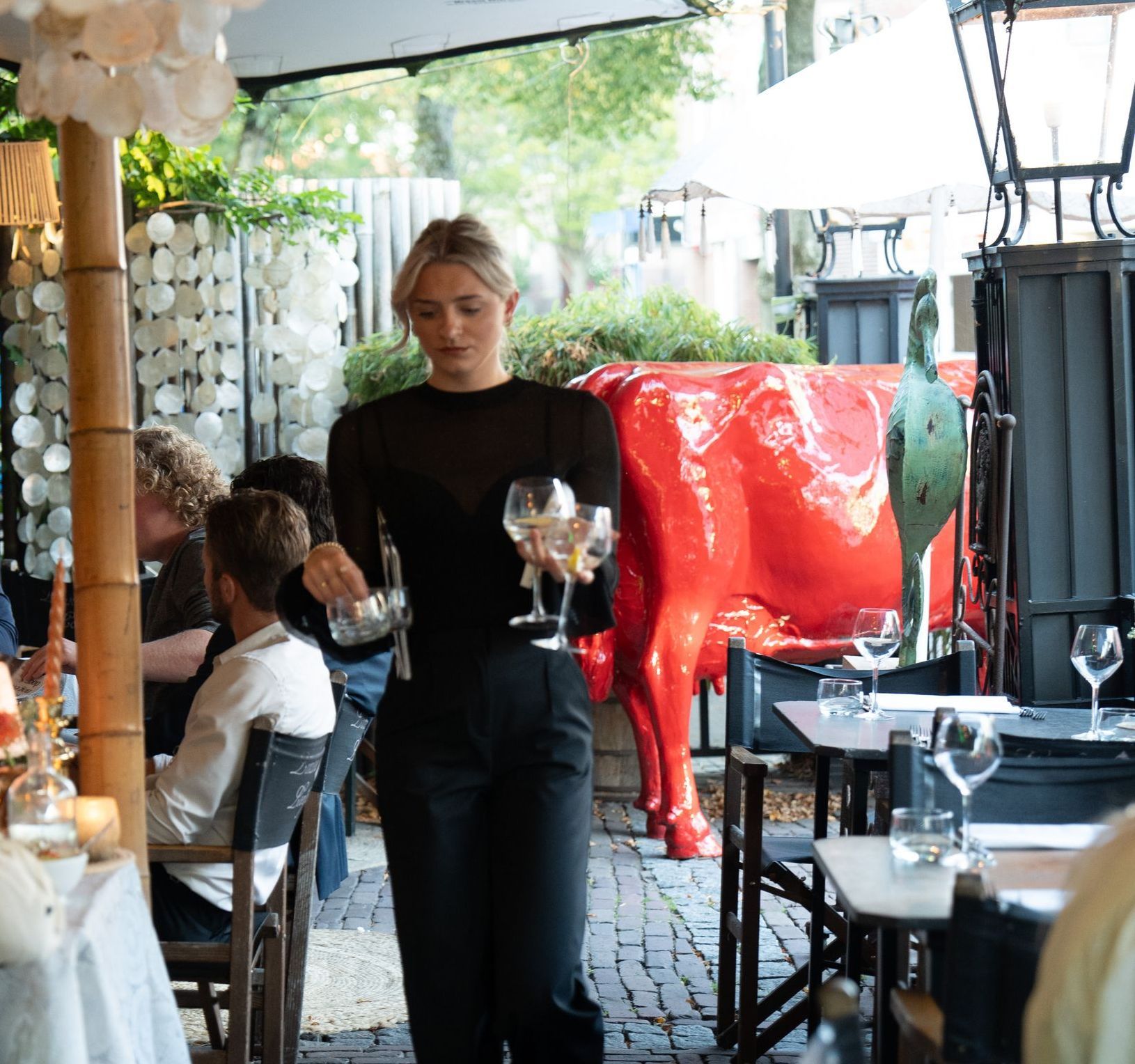 Een vrouw met wijnglazen voor een rood koeienbeeld. Bij Linke Loetje