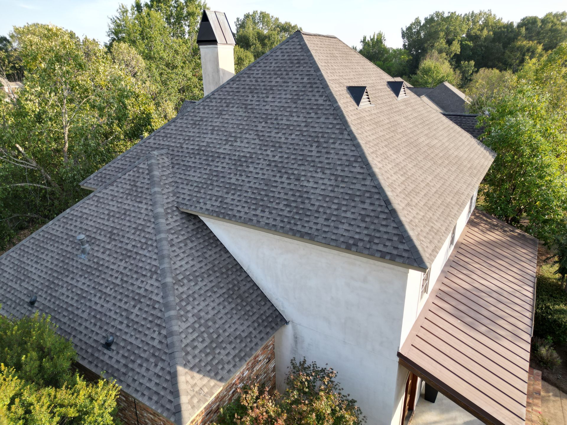 Close-up of a home with a gray shingle roof, chimney, and white stucco exterior surrounded by trees.