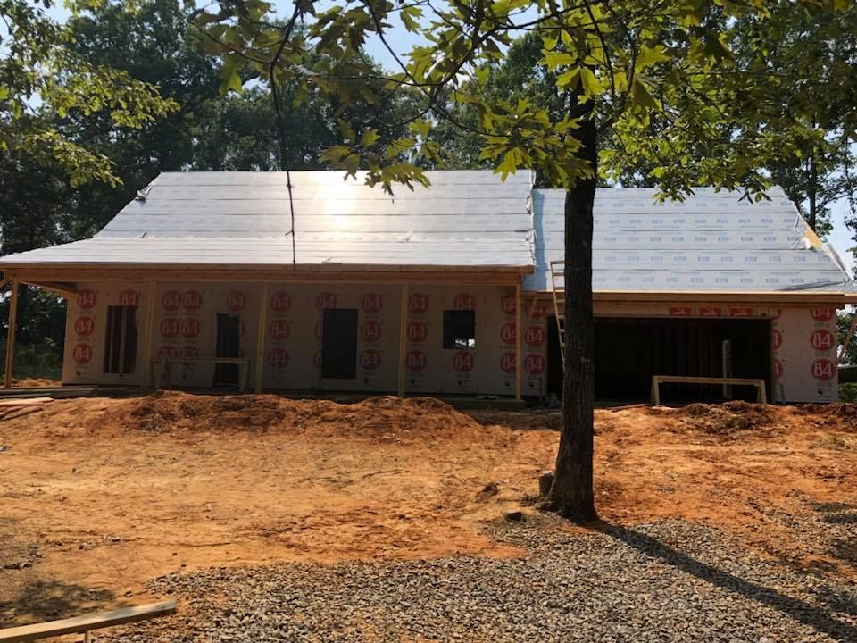 House under construction, exterior view. Building with roof partially completed. Brown dirt and a tree in the foreground.