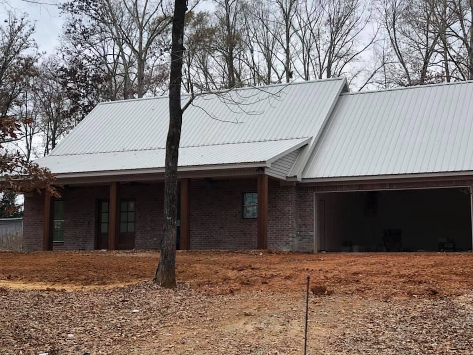 A one-story brick house with a light metal roof and open carport. Brown dirt yard, trees in background.