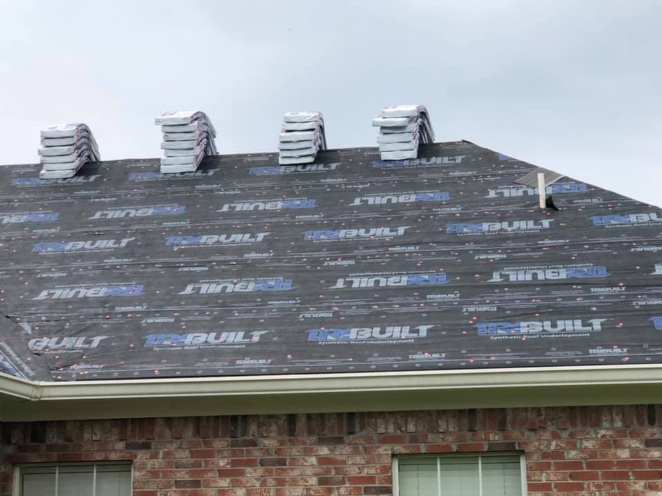 Roof partially covered with underlayment, shingle bundles stacked. Brick house.