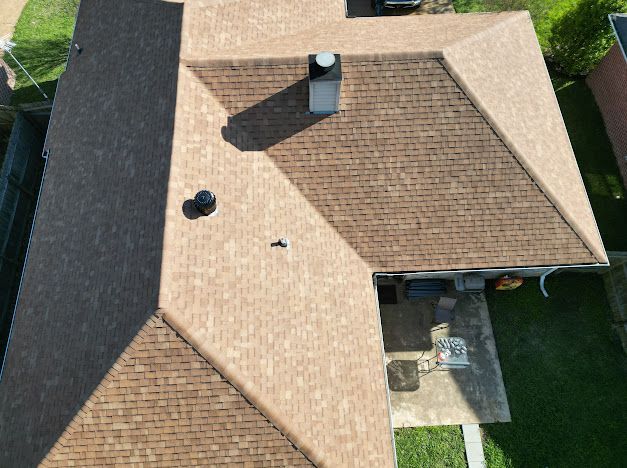 Brown shingle roof of a house, aerial view. Chimney and vents are visible. Sunlight creates shadows.