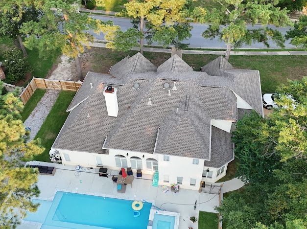 Aerial view of a beige house with a complex brown roof, pool, and surrounding trees.