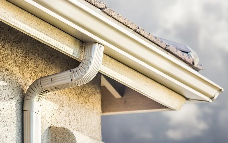 White gutters and downspout on a beige stucco wall, with part of a brown roof visible.