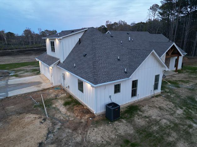 White farmhouse with gray roof, windows, and a black air conditioner, set in a rural landscape.