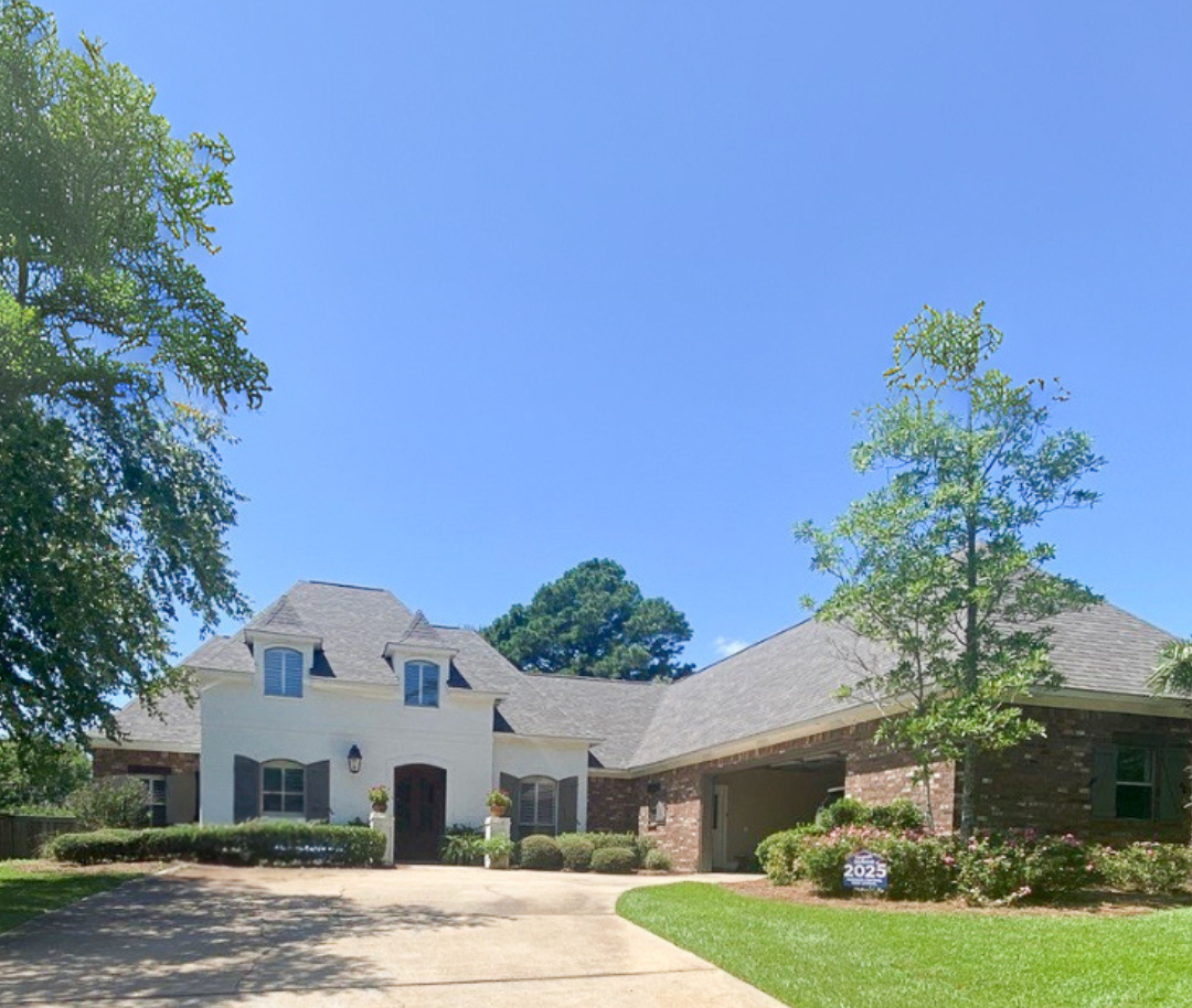 Two-story beige house with a dark gray roof and a long driveway on a sunny day.