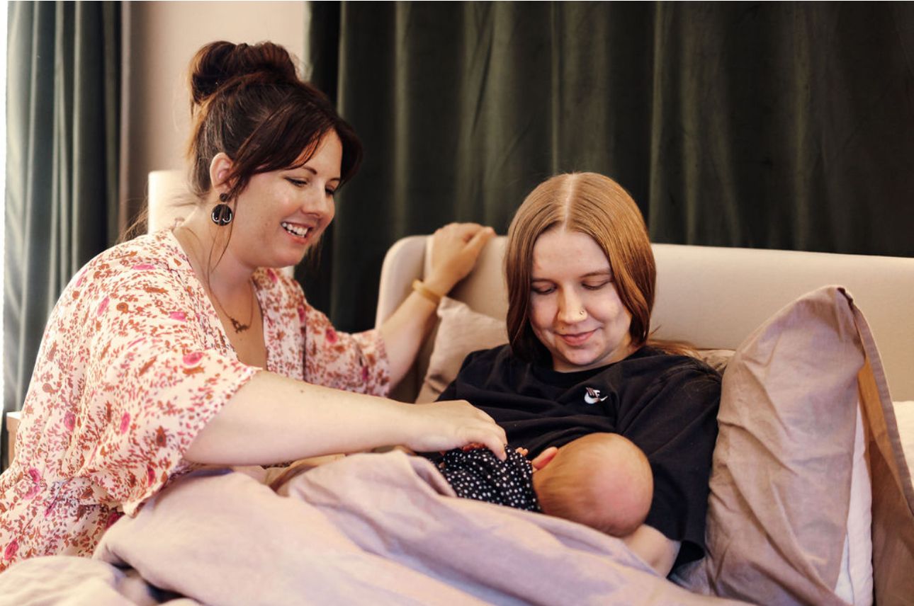 Woman breastfeeding a baby on a bed, another woman beside her, smiling and touching her shoulder.