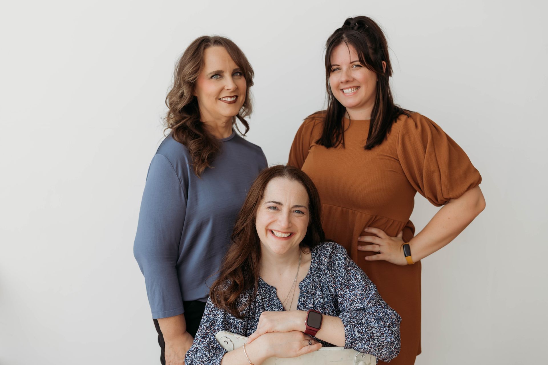 Three women smiling against a white backdrop: one seated, wearing patterned shirt, two standing, one in blue, one in brown.