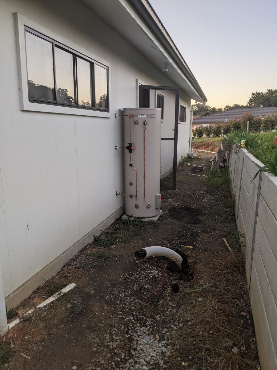 A beige hot water tank attached to a white house exterior, next to a narrow yard with a fence — Robert Coony Electrician in Nambucca Heads, NSW