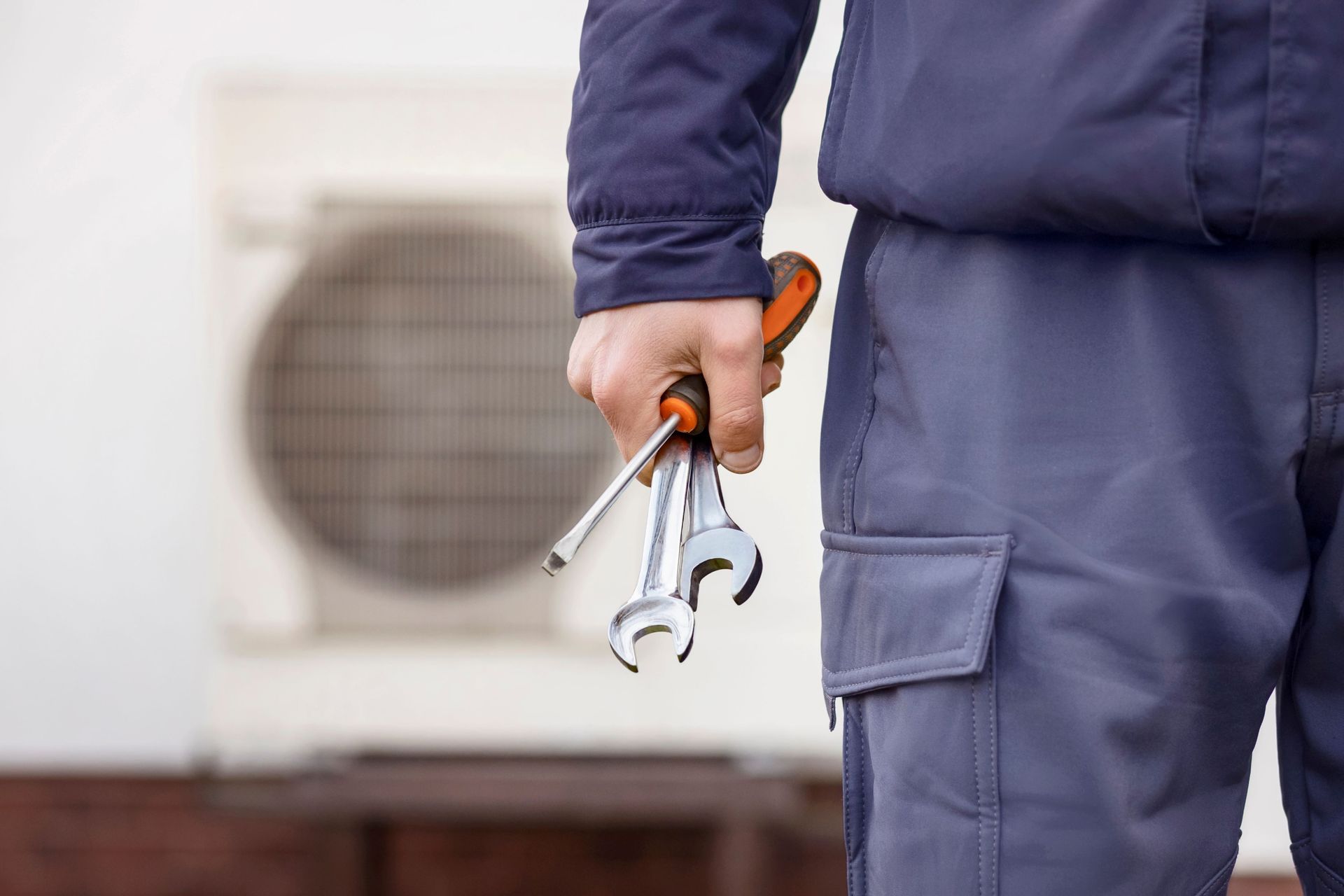 Technician in blue coveralls holding tools beside an outdoor air conditioning unit