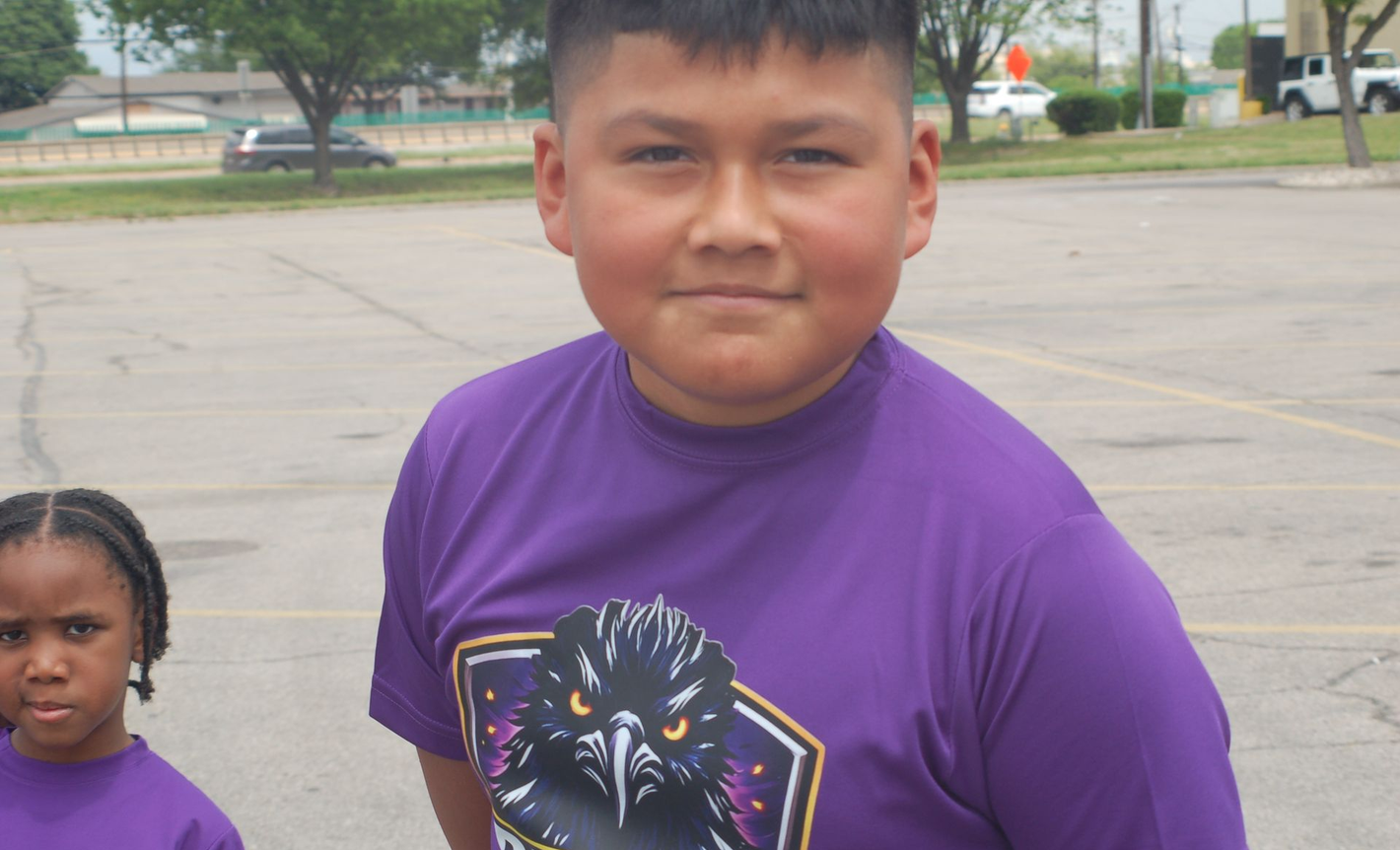 Two children in purple shirts standing outdoors in a parking lot, one centered and looking at the camera.