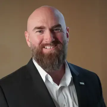 Bald man with beard smiles in suit and white shirt. Brown background.