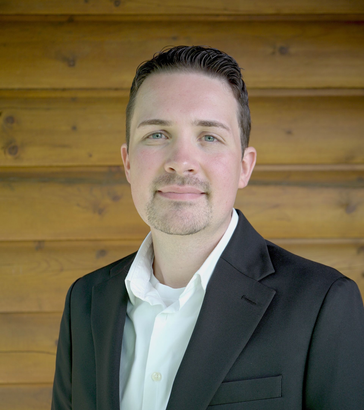 Man with dark hair and goatee smiles in a white shirt and black blazer.