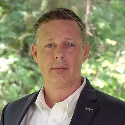 Man in a suit smiles at the camera, with a dark blue jacket, purple tie, and light blue shirt.
