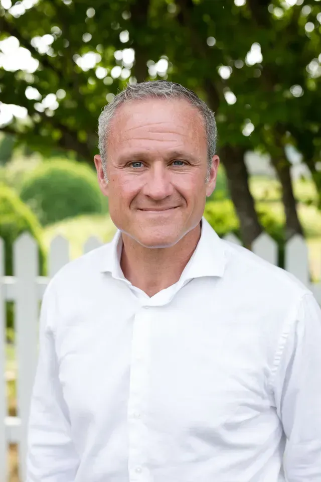 Man in a white shirt smiling in front of a white picket fence and greenery.
