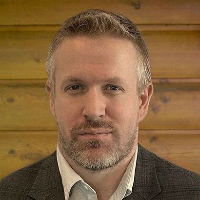 Bald man with beard smiles in suit and white shirt. Brown background.