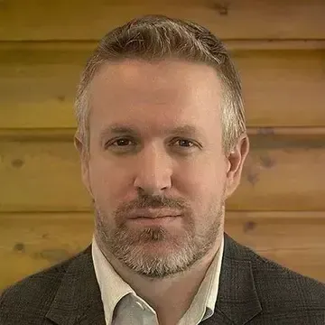 Bald man with beard smiles in suit and white shirt. Brown background.