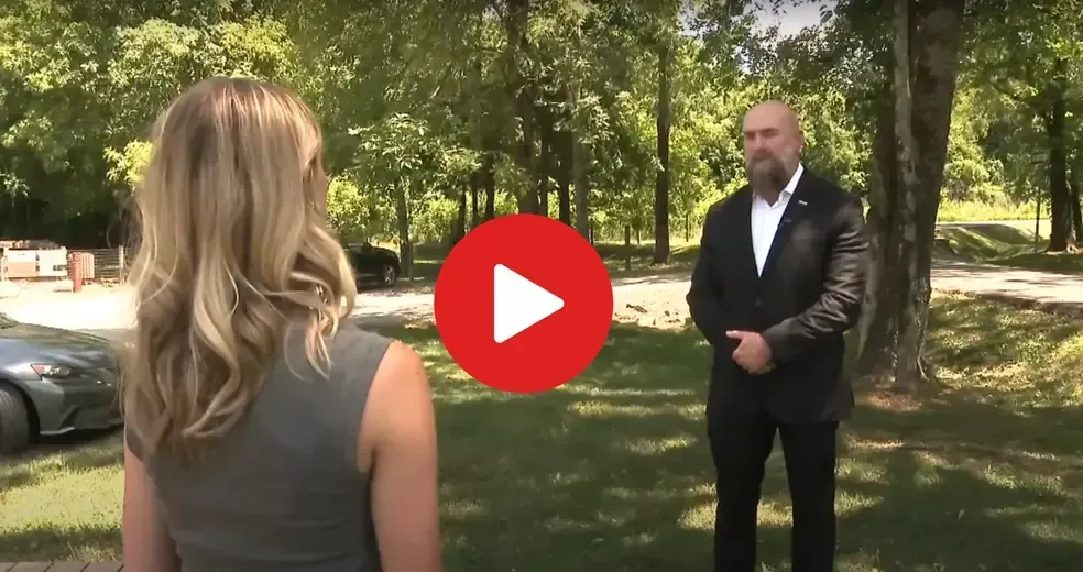 Woman interviewing a man outdoors; trees and a car in the background. The man is wearing a suit.