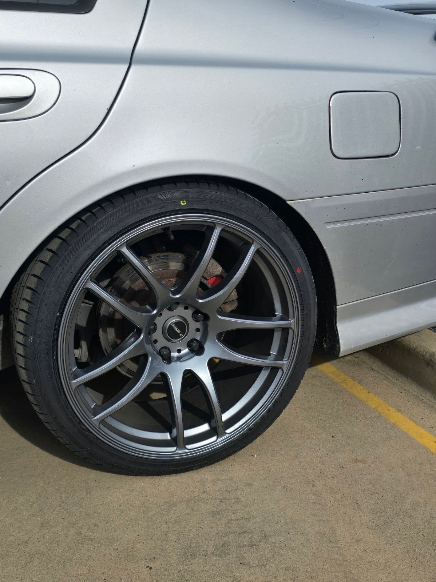 Silver car's rear wheel with a dark gray rim and black tire on a concrete surface. — Peel Valley Tyres in Calala, NSW