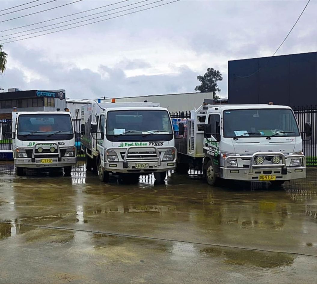Three white work trucks parked on a wet, paved lot under an overcast sky. — Peel Valley Tyres in Taminda, NSW