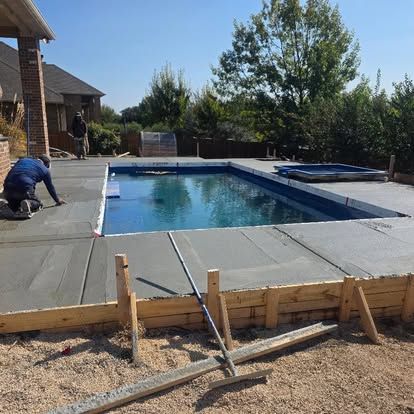 Construction workers pouring concrete around a rectangular pool. Blue water, clear sky, sunny day.