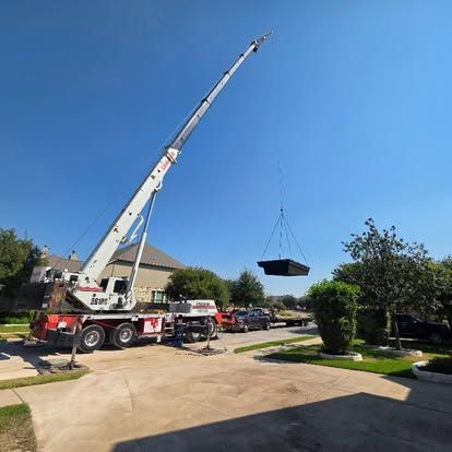 A large crane lifts a rectangular black object over a suburban driveway on a sunny day.