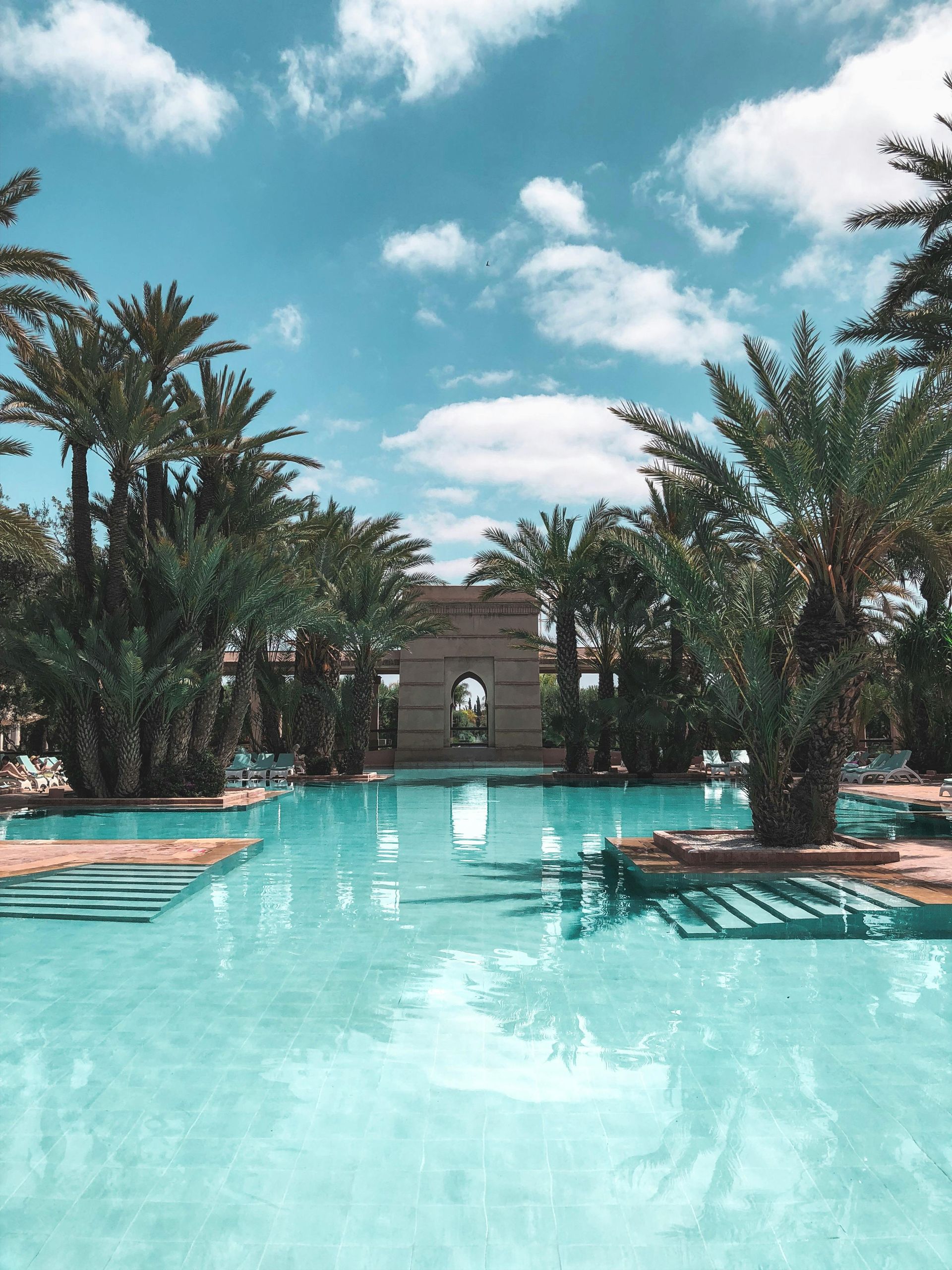 Swimming pool surrounded by palm trees under a bright blue sky. A stone archway is visible in the background.