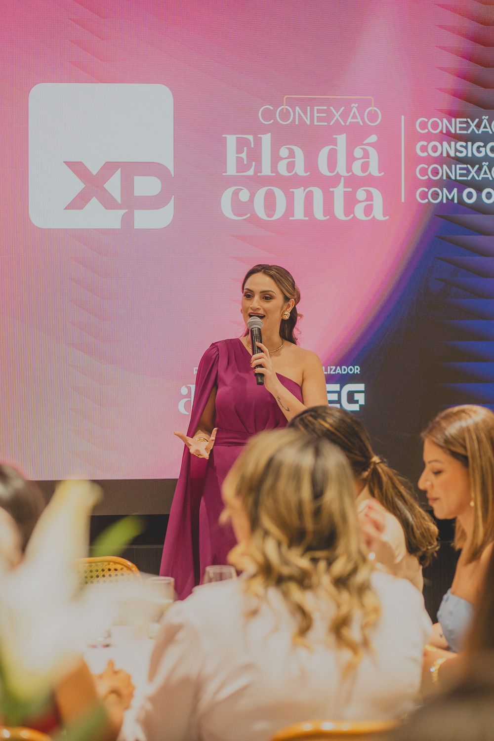 Uma mulher de vestido roxo está parada na frente de um grupo de pessoas segurando um microfone.