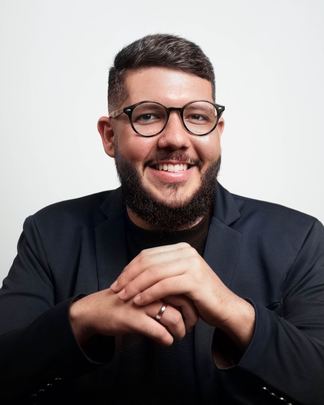 Um homem de óculos e barba sorri com as mãos cruzadas