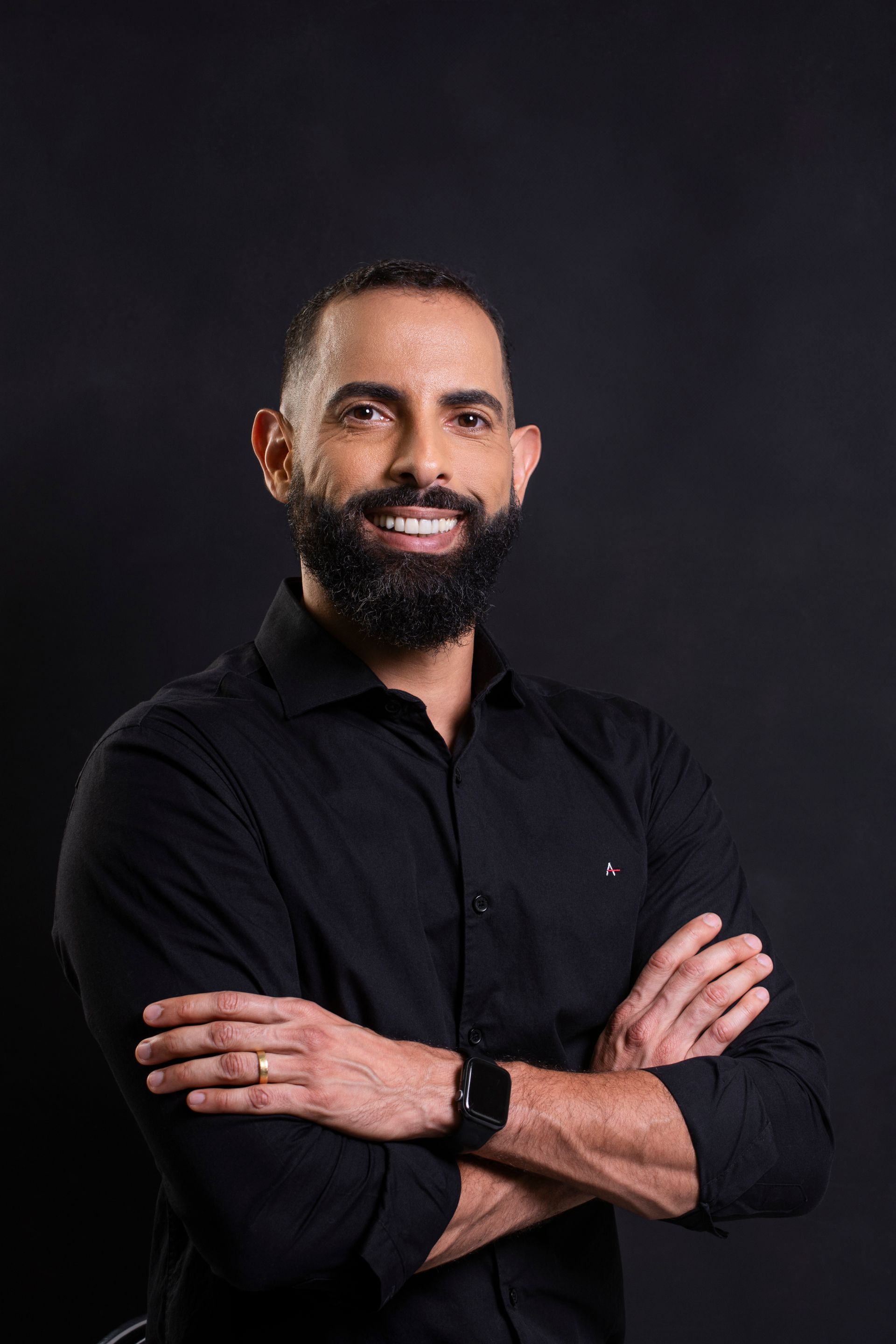 Um homem com barba está vestindo uma camisa preta e está de pé com os braços cruzados.