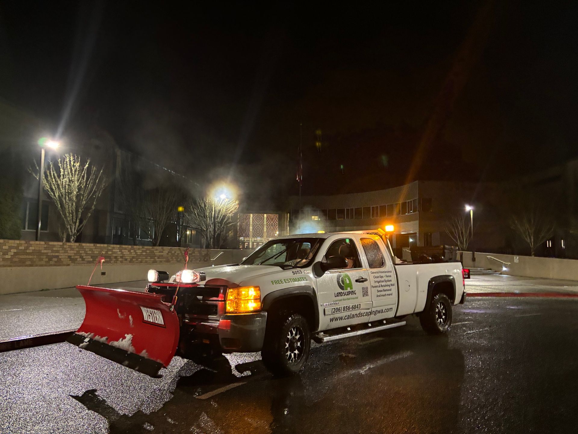 A white truck with a snow plow attached to it is parked on the side of the road at night.