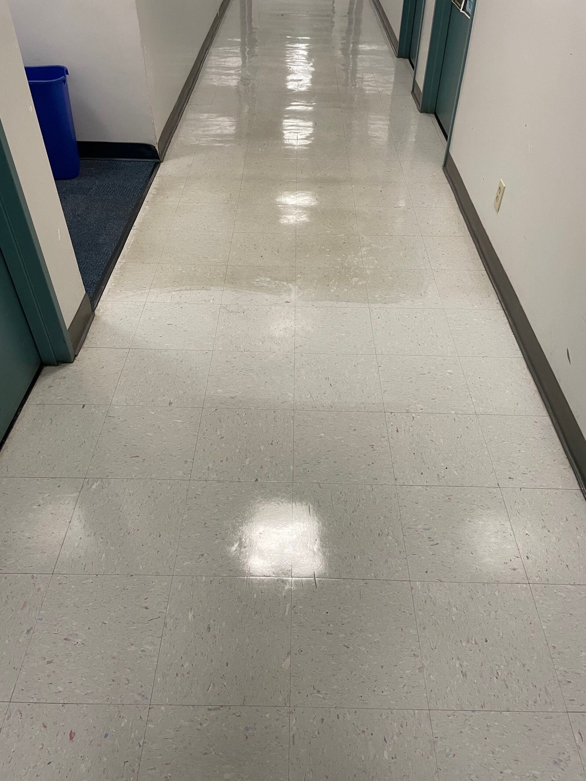 A long hallway with white tiles and a blue trash can in the corner.