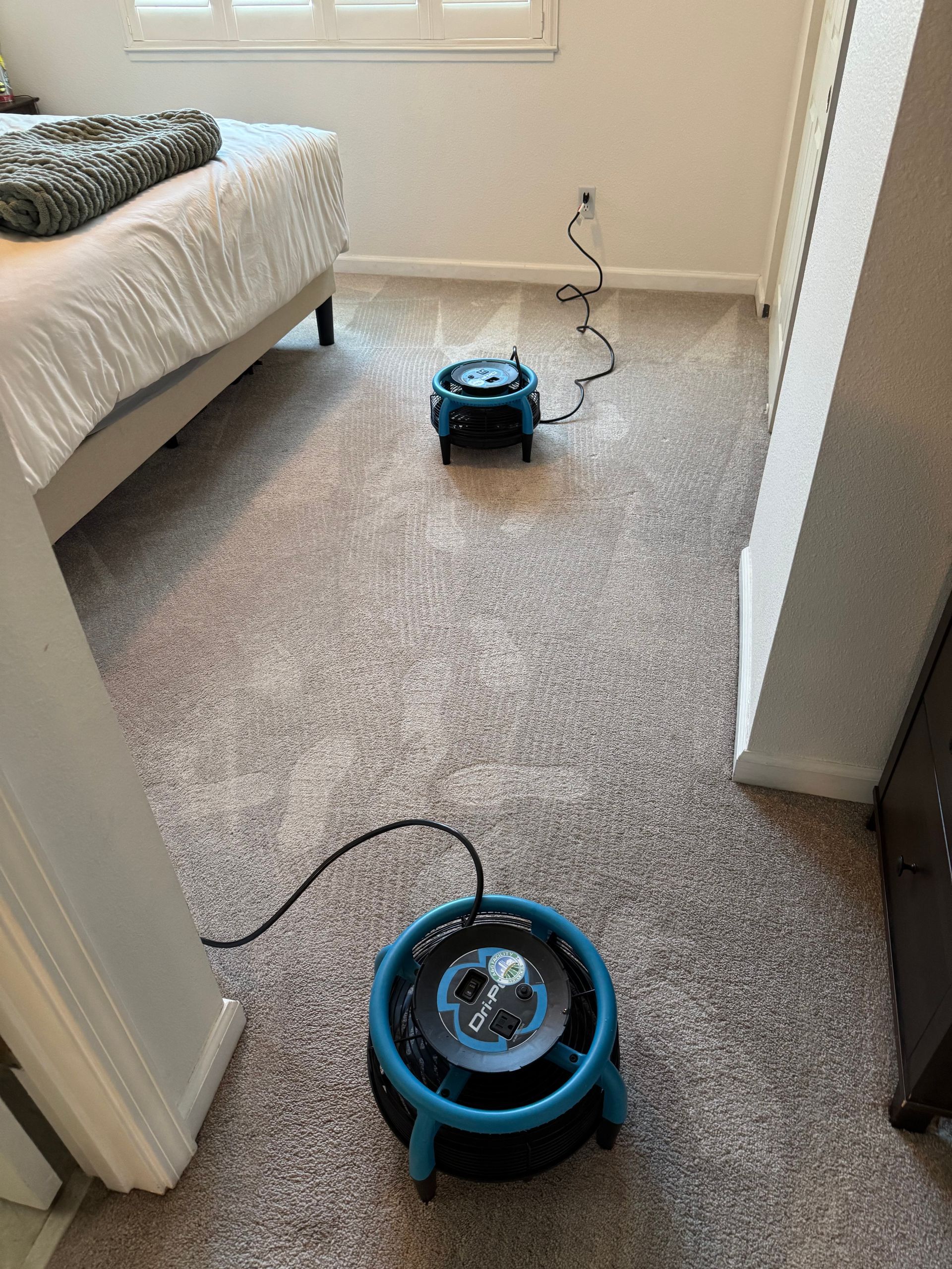 A blue fan is sitting on the floor in a bedroom next to a bed.