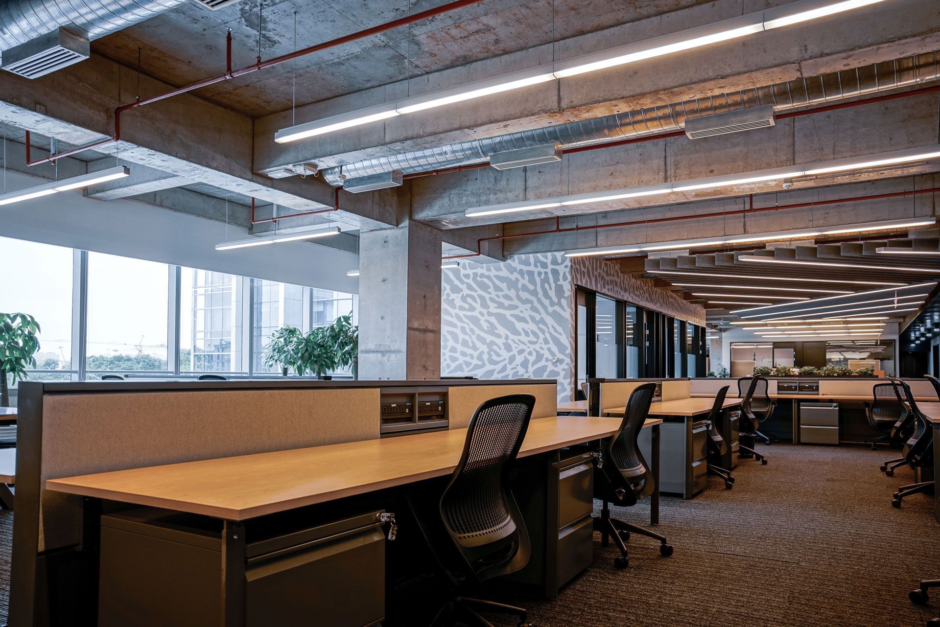 Modern office workspace with desks, chairs, and large windows. Concrete ceiling with linear lighting.
