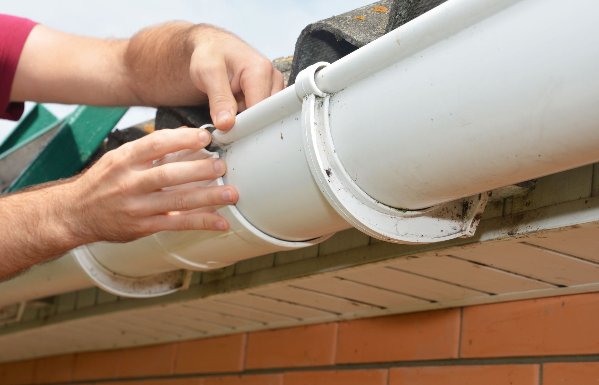Person fixing white gutter on a brick building; hands close up, outdoors.