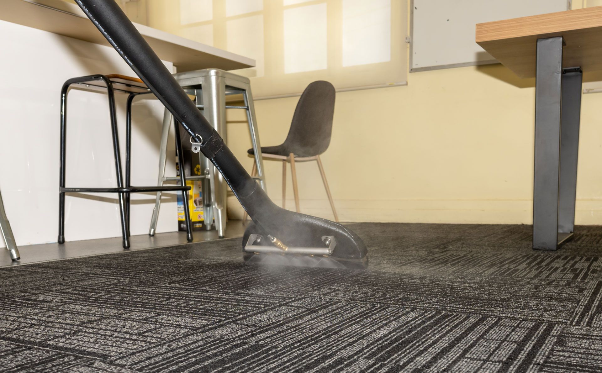 A steam cleaning wand glides over dark gray office carpet tiles, releasing a visible mist of vapor.